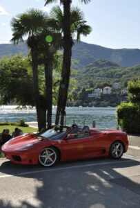 Red Ferrari convertible parked by a scenic lakeside with palm trees and mountain view