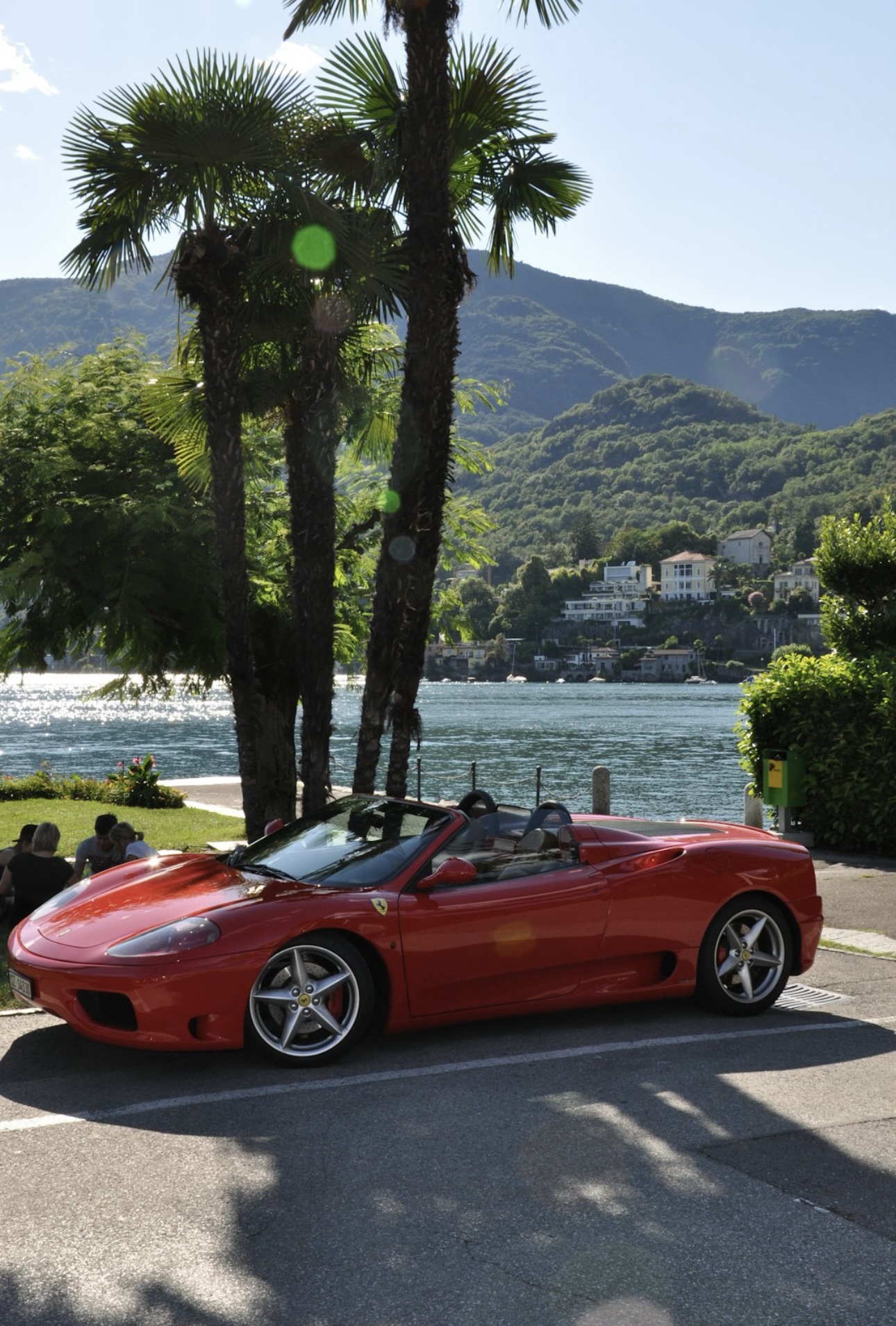 Red Ferrari convertible parked by a scenic lakeside with palm trees and mountain view