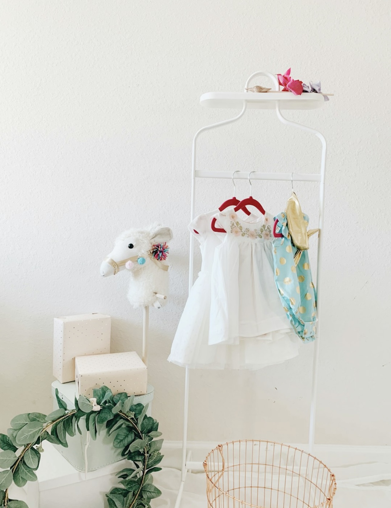 Minimalist baby clothing rack with elegant white and pastel outfits, a stuffed llama toy, gift boxes, and decorative greenery. Aesthetic baby fashion and nursery decor.