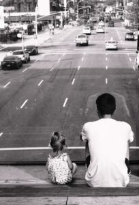 A father and his young daughter sit together on a wooden bench, overlooking a busy city street. The father, wearing a white t-shirt, and the daughter, dressed in a patterned outfit, share a quiet moment of reflection. A black and white photograph capturing the deep bond between a father and child.