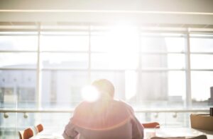 Back view of a man sitting at a desk in a bright modern office, facing large windows with sunlight streaming in—symbolizing workplace stress, isolation, and mobbing.